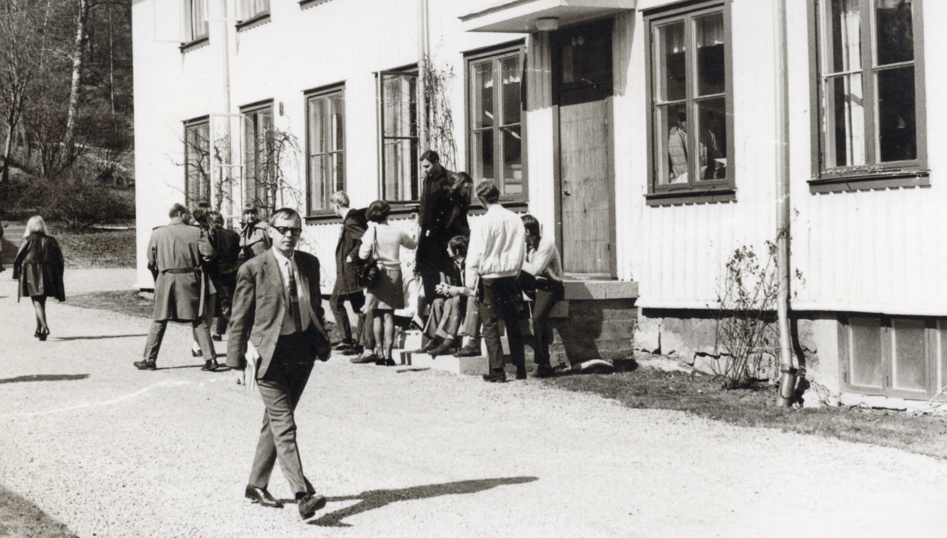 Svartvitt kort med vuxna deltagare framför skolbyggnad. Lärare Åke Forsman i kavaj och slips.