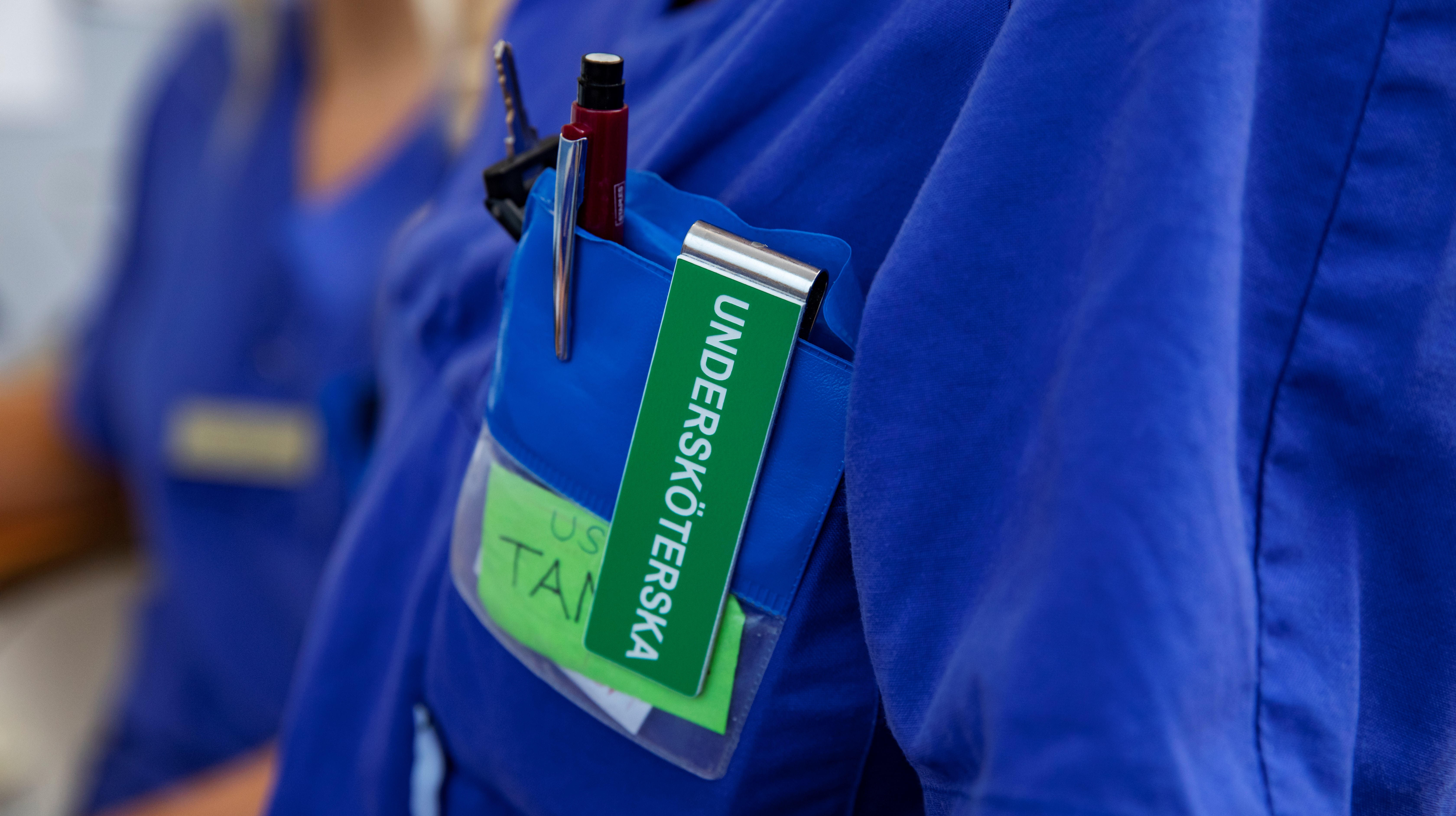 En blå tröja där en clip med texten "undersköterska" och en penna hänger i bröstfickan.